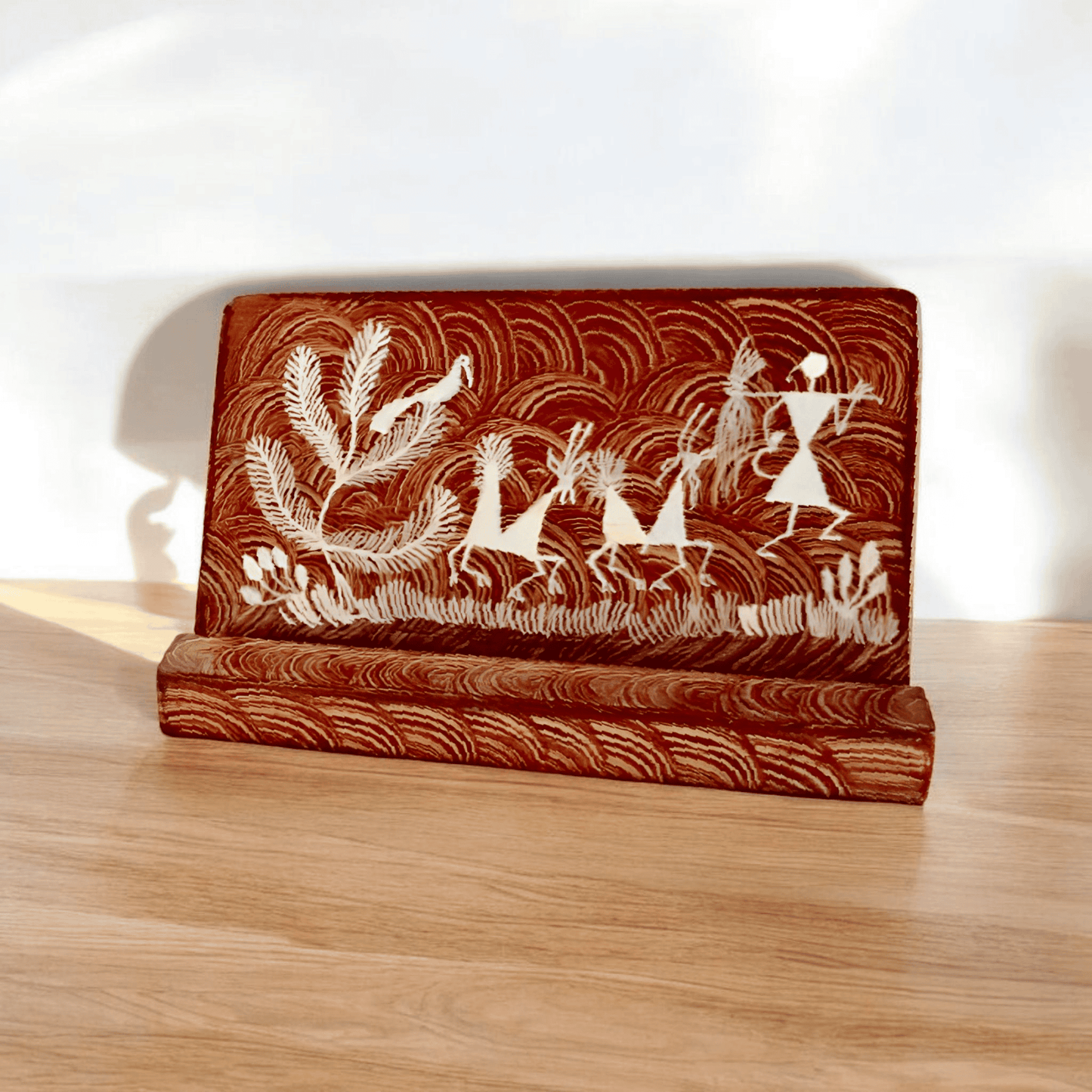 A wooden phone stand with hand-painted Warli tribal patterns in brown and white colors, placed on a wooden surface.
