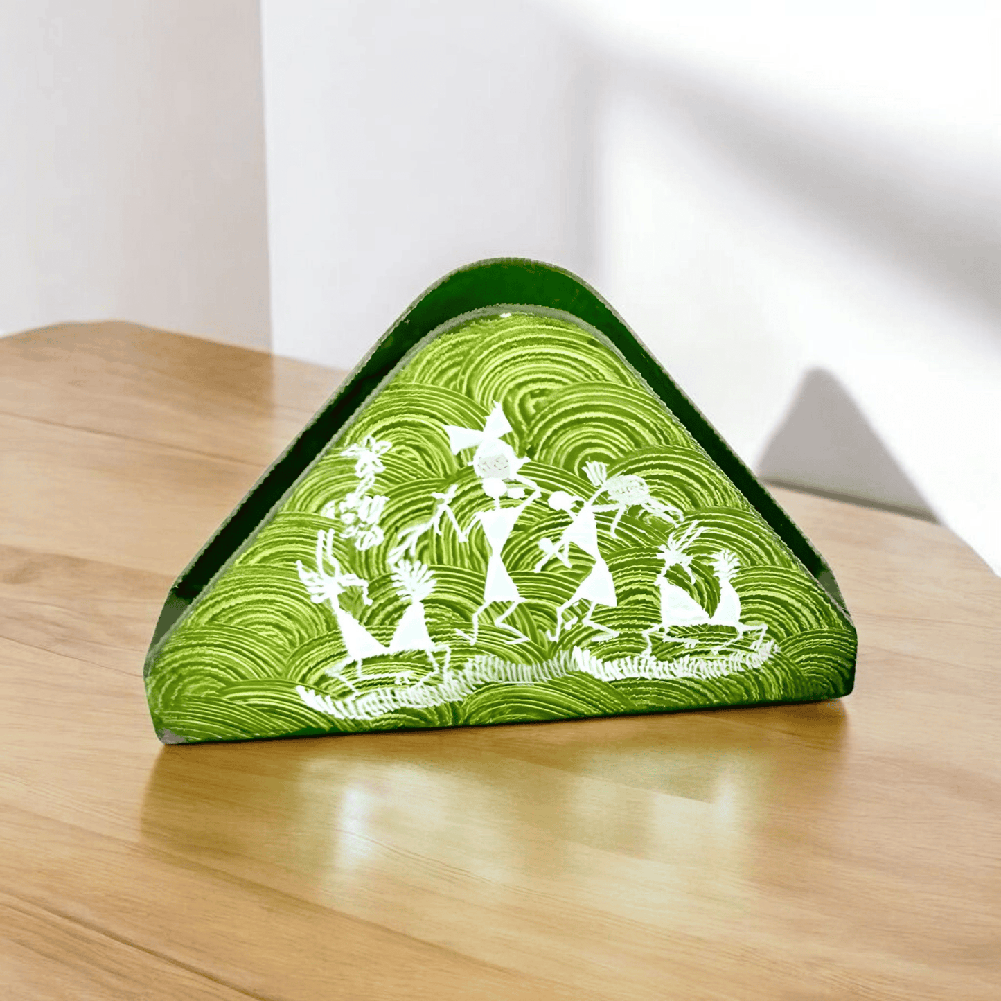 A triangular wooden napkin holder with Warli tribal artwork, predominantly green with white patterns, placed on a wooden surface.