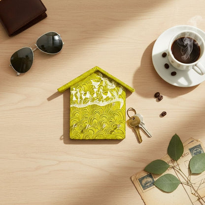 Green house-shaped keychain holder  on a wooden surface with a cup of coffee, keys, and sunglasses.